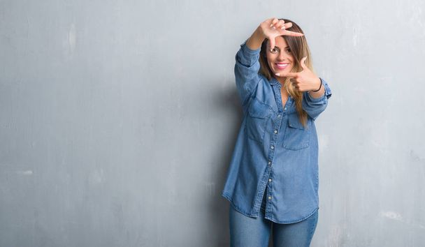 Молодая взрослая женщина за гранж серой стеной в джинсовой одежде улыбается делая рамку с руками и пальцами с счастливым лицом. Концепция творчества и фотографии
. - Фото, изображение
