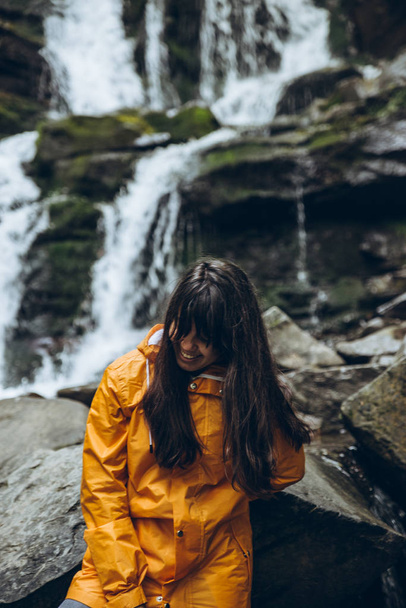 滝の背景に黄色のレインコートに笑顔の女性。ハイキングのコンセプト - 写真・画像