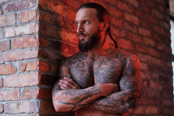 Elegante hombre barbudo con el cuerpo musculoso y tatuado apoyado en una pared con los brazos cruzados y mirando por la ventana
. - Foto, Imagen