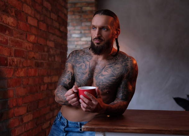 刺青ボディとひげを生やした男性ロフトのインテリアの部屋のテーブルの上にもたれながら一杯のコーヒーを保持しています。. - 写真・画像