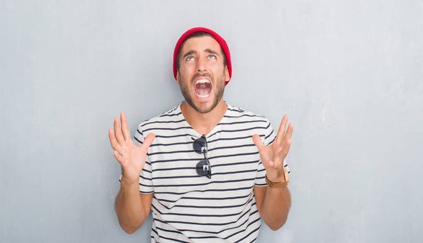 Guapo joven hipster hombre sobre gris pared grunge con camiseta azul marino y gorra de lana loco y loco gritando y gritando con expresión agresiva y los brazos levantados. Concepto de frustración
. - Foto, Imagen