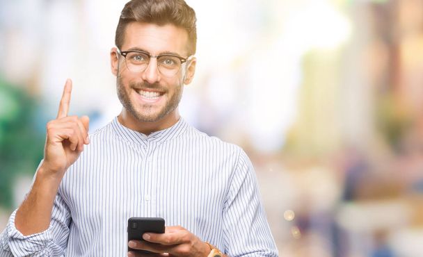 Młody przystojny mężczyzna SMS-y za pomocą smartfona na białym tle zaskoczony z pomysłu lub pytanie wskazując palcem z szczęśliwy twarz, numer jeden - Zdjęcie, obraz