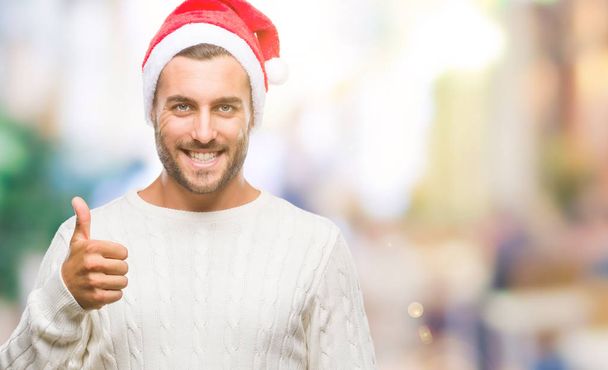 Jonge knappe man Kerstman hoed dragen over geïsoleerde achtergrond gelukkig duimschroef opwaarts gebaar met de hand te doen. Goedkeuring van expressie kijken naar de camera met tonen van succes. - Foto, afbeelding