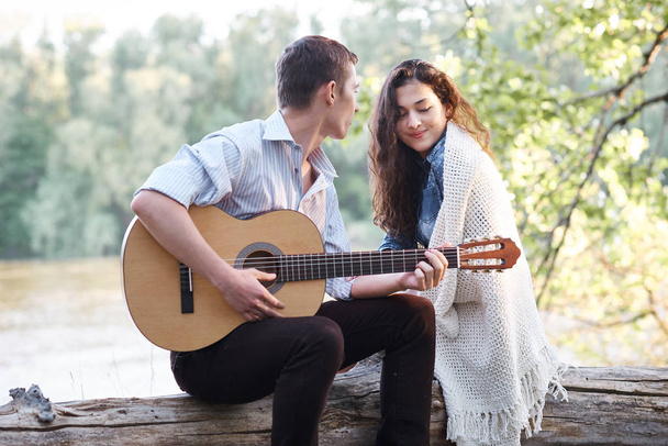 young couple sitting on a log by the river and playing guitar, summer nature, bright sunlight, shadows and green leaves, romantic feelings - Photo, Image