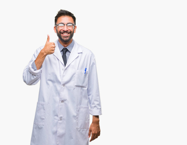 Erwachsener hispanischer Wissenschaftler oder Arzt, der einen weißen Mantel über isoliertem Hintergrund trägt und mit der Hand fröhlich Daumen hoch gestikuliert. zustimmender Ausdruck, der mit Erfolg in die Kamera blickt. - Foto, Bild
