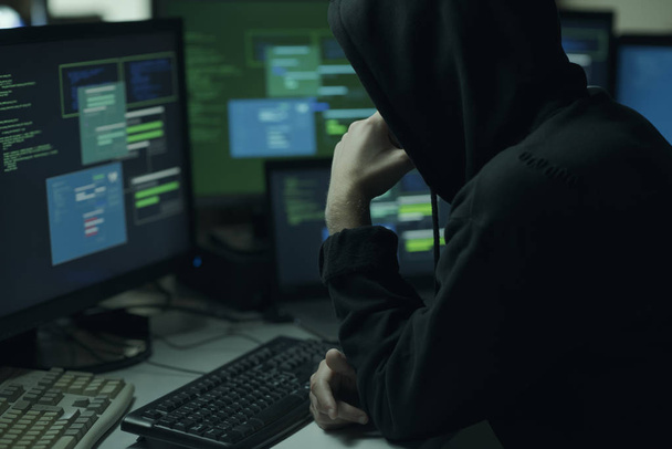 Young hacker with a hoodie thinking with hand on chin and working with computers, technology and cybercrime concept - Foto, afbeelding