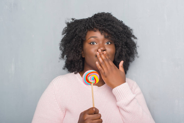 Fiatal afrikai-amerikai nő evés nyalóka cukorka terjed száj kézzel döbbenve, hiba, kifejezés, a félelem, szégyen, szürke grunge falon ijedt csendben, titkos koncepció - Fotó, kép