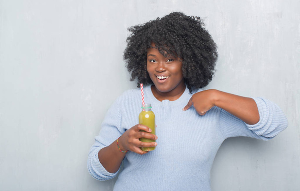 Giovane donna afroamericana sopra grigio grunge parete bere detox succo verde con faccia a sorpresa puntando il dito verso se stesso
 - Foto, immagini