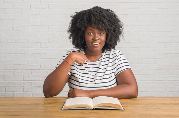 junge afrikanisch-amerikanische Frau sitzt am Tisch und liest ein Buch mit überraschendem Gesicht, das mit dem Finger auf sich selbst zeigt - Foto, Bild
