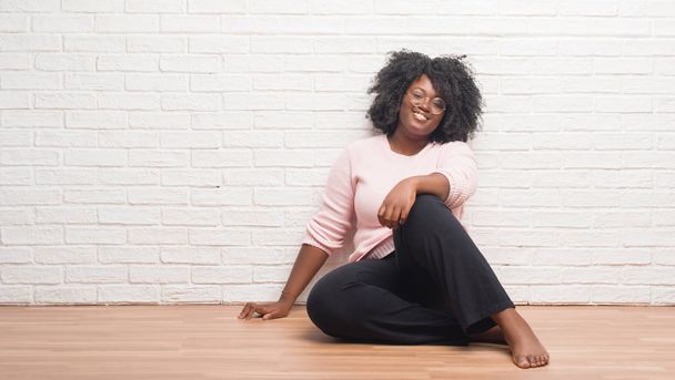 Νεαρός αφρικανική αμερικανική γυναίκα που κάθεται στο πάτωμα στο σπίτι με μια ευτυχισμένη και δροσερό χαμόγελο στο πρόσωπο. Τυχερό πρόσωπο. - Φωτογραφία, εικόνα