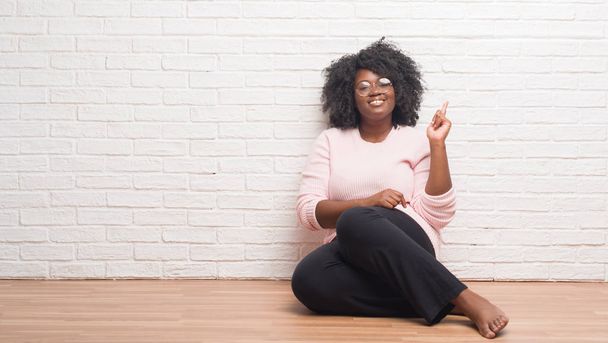 Jonge african american vrouw zittend op de vloer thuis met een grote glimlach op het gezicht, met de hand en de vinger te wijzen naar de kant te kijken naar de camera. - Foto, afbeelding