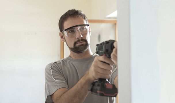 Professional repairman with protective goggles, he is using a drill and doing a home renovation - 写真・画像