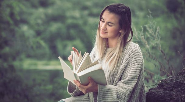 girl in a sweater reads a book against a background of nature, a concept of leisure and a hobby - Photo, image