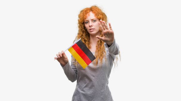 Jeune femme rousse tenant le drapeau de l'Allemagne avec la main ouverte faisant stop signe avec expression sérieuse et confiante, geste de défense
 - Photo, image