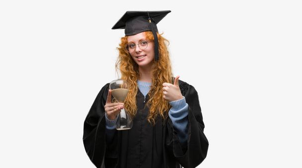 Jovem estudante ruiva mulher vestindo uniforme graduado segurando ampulheta feliz com grande sorriso fazendo sinal ok, polegar para cima com os dedos, excelente sinal
 - Foto, Imagem
