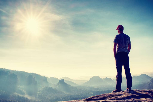 Высокий человек в наружной одежде стоит один на вершине скалы. Турист смотрит холмистый лесной пейзаж в утреннем тумане
. - Фото, изображение