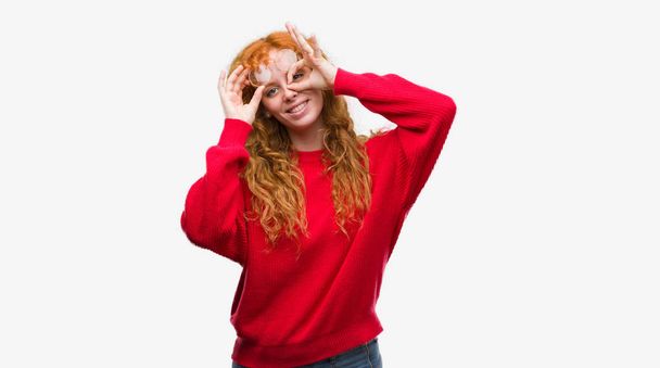 Jonge roodharige vrouw dragen rode trui doen ok gebaar met hand glimlachen, oog kijken door vingers met blij gezicht. - Foto, afbeelding