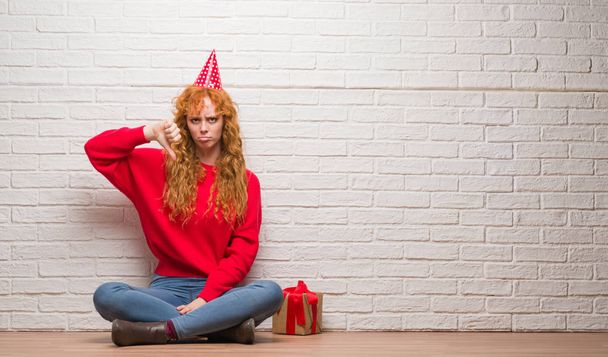 怒った顔、ダウン親指で嫌悪感を示すマイナス記号、拒絶反応の概念を身に着けている誕生日帽子壁のレンガの上に座っている赤毛の若い女性 - 写真・画像