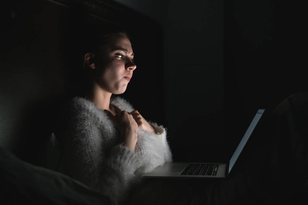young scared girl sitting in bed late at night in the dark watching a horror film on her laptop - Fotografie, Obrázek