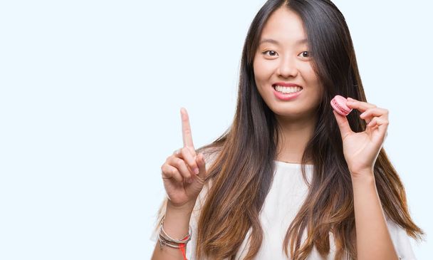 Young asian woman eating pink macaron sweet over isolated background surprised with an idea or question pointing finger with happy face, number one - Foto, Imagen