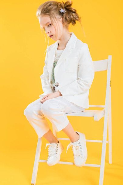 A teenage girl in white clothes is sitting on a chair on a yellow background. - Fotografie, Obrázek