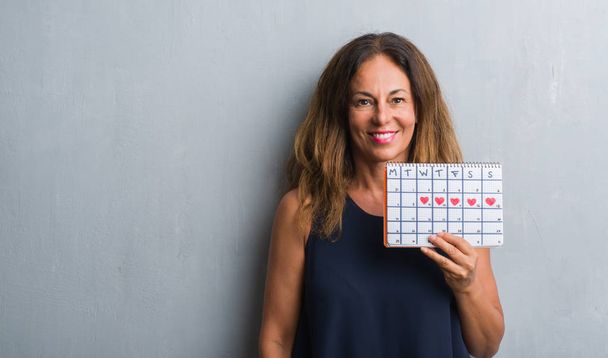 Middelbare leeftijd Latino vrouw over grijs grunge muur periode kalender met een blij gezicht permanent en lachend met een vertrouwen glimlach weergeven tanden houden - Foto, afbeelding