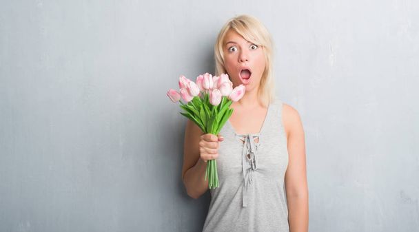 Femme adulte caucasienne sur un mur gris grunge tenant des fleurs roses effrayées en état de choc avec un visage surprise, effrayées et excitées par l'expression de la peur
 - Photo, image