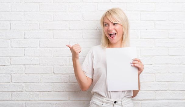 Mujer caucásica adulta sobre pared de ladrillo blanco sosteniendo hoja de papel en blanco señalando y mostrando con el pulgar hacia el lado con la cara feliz sonriendo
 - Foto, imagen