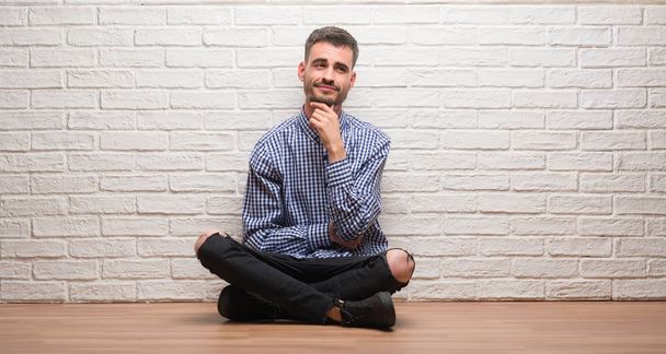 Νεαρός ενήλικος άνδρας κάθεται πάνω από το λευκό τοίχο με το χέρι στο πηγούνι σκέψης σχετικά με την ερώτηση, συλλογισμένος έκφραση. Χαμογελώντας με το στοχαστικό πρόσωπο. Έννοια της αμφιβολίας. - Φωτογραφία, εικόνα