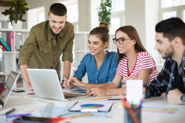 Jovens empresários positivos trabalhando alegremente no laptop juntos. Grupo de homens e mulheres sorridentes que passam o tempo no trabalho no escritório acolhedor moderno
 - Foto, Imagem