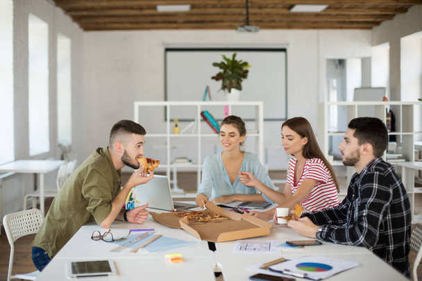 Група молодих творчих людей їсть піцу разом в очікуванні роботи в сучасному затишному офісі
 - Фото, зображення