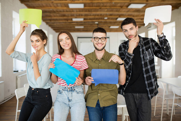 Group of young cheerful people holding colorful papers messages icons in hands happily looking in camera spending time at work in modern office - Photo, Image