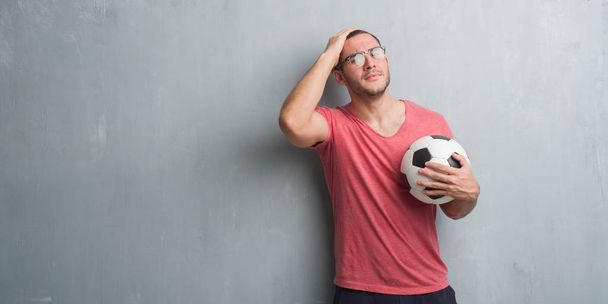 Young caucasian man over grey grunge wall holding soccer football ball stressed with hand on head, shocked with shame and surprise face, angry and frustrated. Fear and upset for mistake. - Photo, Image