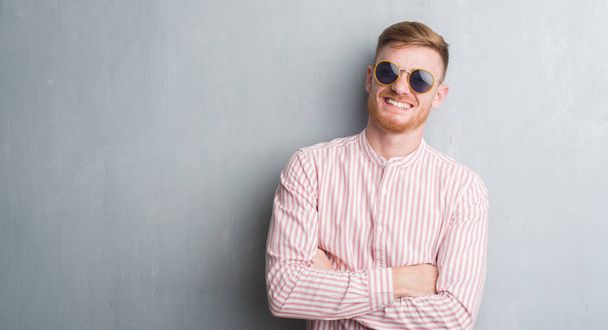Молодий рудий чоловік над сірою гранжевою стіною в ретро сонцезахисних окулярах з щасливим обличчям, що стоїть і посміхається з впевненою посмішкою, показуючи зуби
 - Фото, зображення