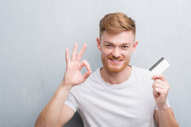 Homem ruivo jovem sobre parede grunge cinza segurando cartão de crédito fazendo sinal ok com os dedos, excelente símbolo
 - Foto, Imagem