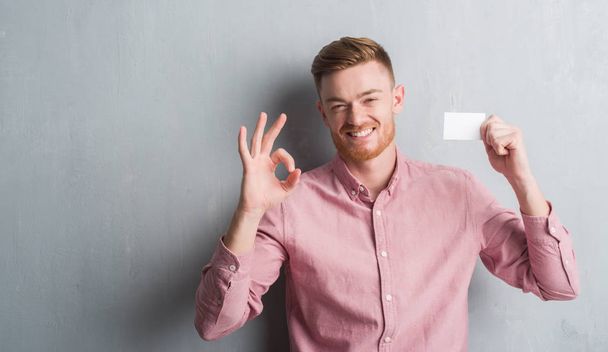 Genç Kızıl saçlı adam parmak, mükemmel sembolü ile ok işareti yapıyor boş ziyaret Kart tutan gri grunge duvarın üzerinden - Fotoğraf, Görsel