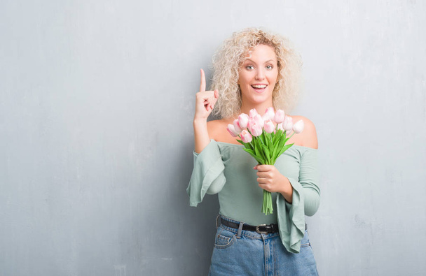 Jonge blonde vrouw over grunge grijze achtergrond met bloemen verrast met een idee of vraag wijzende vinger met blij gezicht, nummer één - Foto, afbeelding