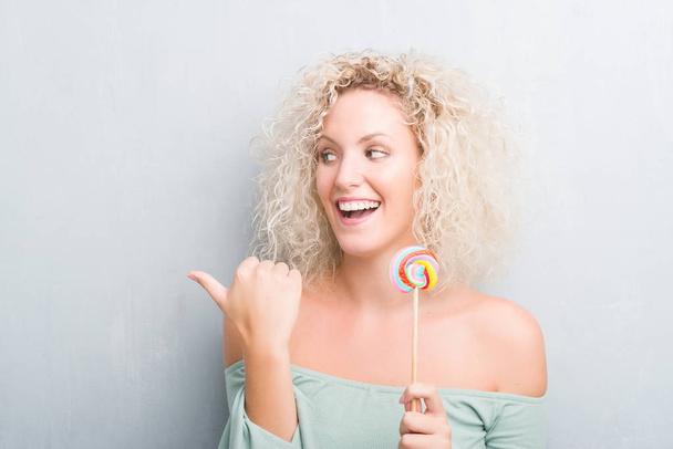 Młoda blond kobieta streszczenie szary mur jedzenie candy lollipop wskazując i Wyświetlono z kciukiem do boku z szczęśliwy twarz uśmiechający się - Zdjęcie, obraz