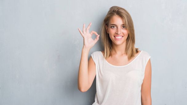 Beautiful young woman standing over grunge grey wall doing ok sign with fingers, excellent symbol - Photo, Image