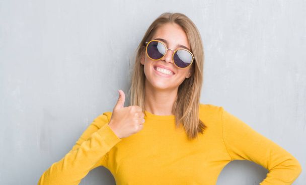 Kaunis nuori nainen seisoo grunge harmaa seinä yllään retro aurinkolasit onnellinen iso hymy tekee ok merkki, peukalo ylös sormilla, erinomainen merkki
 - Valokuva, kuva