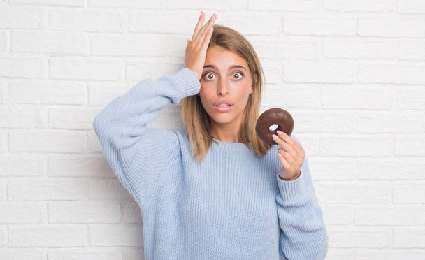 Mooie jonge vrouw over witte bakstenen muur eten chocolade donut benadrukt met de hand op het hoofd, geschokt met schaamte en verrassing gezicht, boos en gefrustreerd. Angst en boos voor fout. - Foto, afbeelding