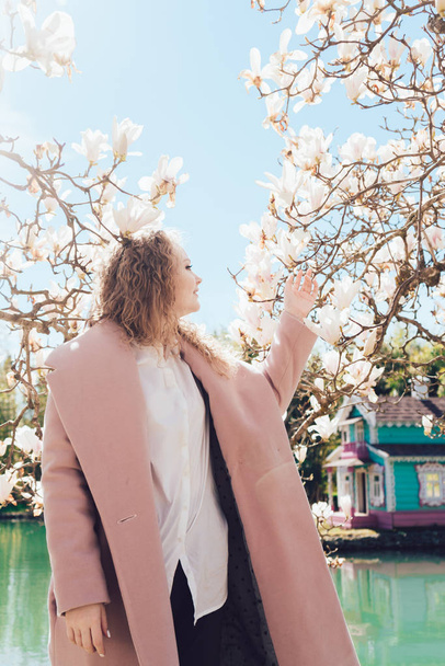 a luxurious curly woman in a trendy coat enjoying nature in the garden, a scented magnolia - Photo, Image