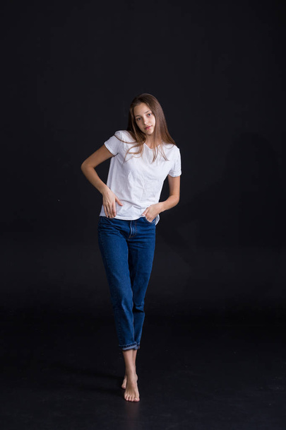 Beautiful girl with brown hair and blue eyes iin white t-shirt and blue jeans solated on a dark background - Foto, afbeelding
