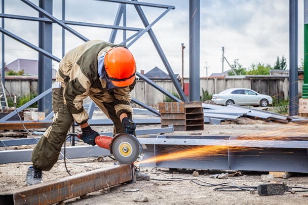 A strong  man welder in brown uniform, a construction helmet and welders leathers, grinder metal an angle grinder  at the construction site, orange sparks fly to the sides - Photo, Image