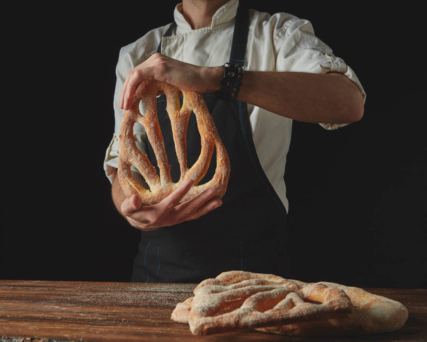 On a dark background and on a background of a wooden brown table, men's hands hold freshly baked bread fougas - Фото, изображение