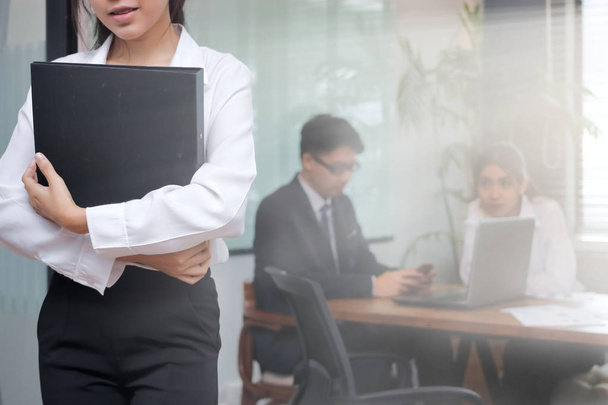 Leadership business woman concept. Confident young Asian businesswoman with ring binder standing against her colleague in office background. - Фото, зображення