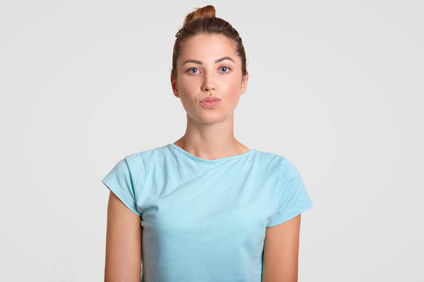 Horizontal shot of pleasant looking woman with healthy skin, keeps lips rounded, dressed in casual blue light t shirt, isolated over white background. People, facial expressions and skin care concept - Fotoğraf, Görsel