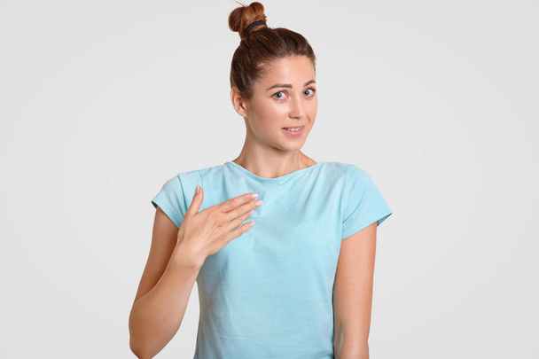 Unsure beautiful woman keeps hand on chest, asking why she was chosen, stands against white studio wall, wears casul t shirt, isolated over white background. People doubt and shyness concept. - Fotoğraf, Görsel