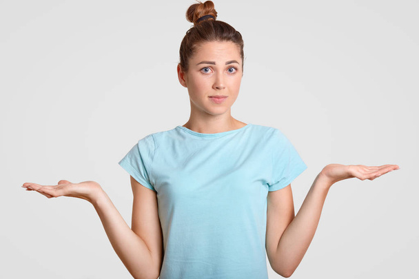 Hesitant young woman shrugs shoulders with uncertainy, cant make decision, clasps palms, looks clueless at camera, dressed in blue t shirt, isolated over white background. So, what to do now? - 写真・画像