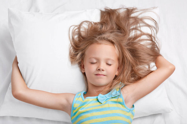 Indoor shot of adorable small female kid with uncombed light hair, keeps hands under soft pillow, dressed in pyjamass, poses in comfortable bed alone. Children, night rest and lifestyle concept - Φωτογραφία, εικόνα